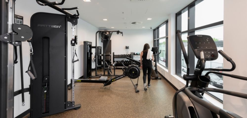 A woman walks through a bright modern gym lined with strength machines and cardio equipment, creating a clean, focused workout atmosphere.
