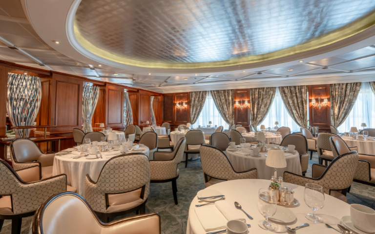 An elegant dining room with cream-clothed tables, patterned chairs, tall curtained windows and a glowing oval ceiling onboard Regatta.