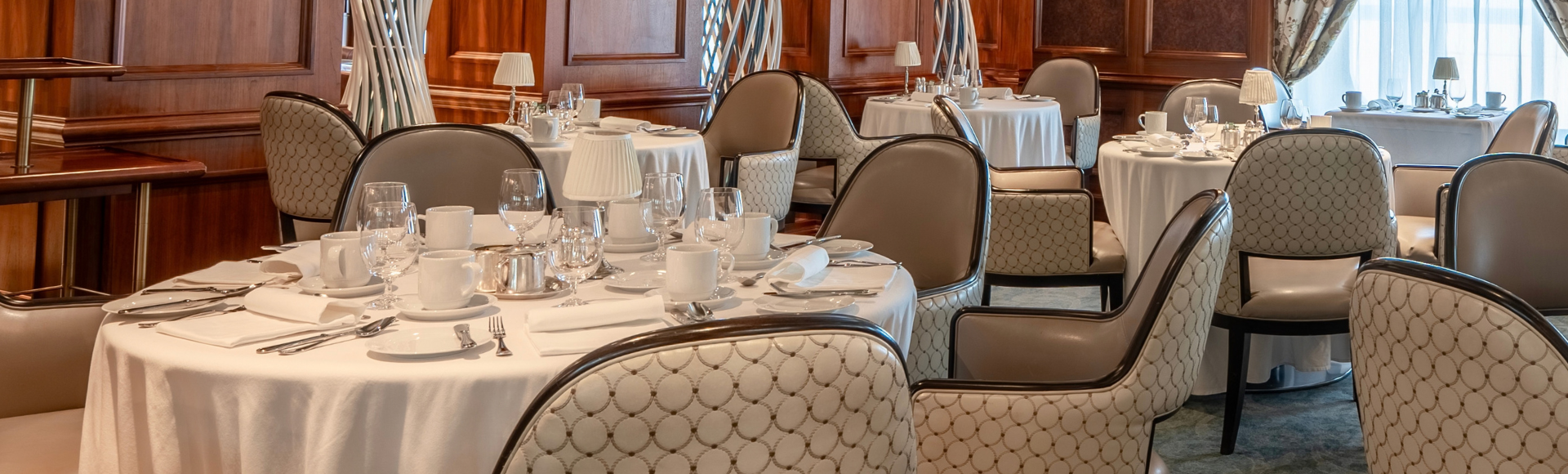 An elegant dining room with cream-clothed tables, patterned chairs, tall curtained windows and a glowing oval ceiling onboard Regatta.