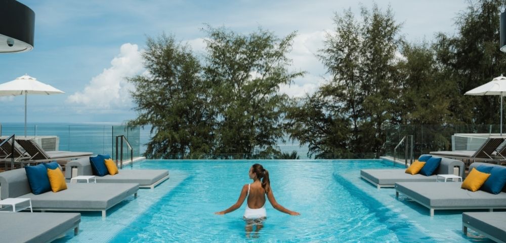 A person in a white swimsuit stands in an infinity pool on a sunny day, with lounge chairs and umbrellas visible around the pool's edge, and lush trees and the ocean in the background.