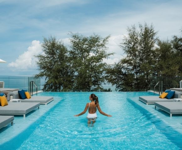 A person in a white swimsuit stands in an infinity pool on a sunny day, with lounge chairs and umbrellas visible around the pool's edge, and lush trees and the ocean in the background.