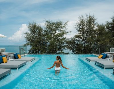 A person in a white swimsuit stands in an infinity pool on a sunny day, with lounge chairs and umbrellas visible around the pool's edge, and lush trees and the ocean in the background.