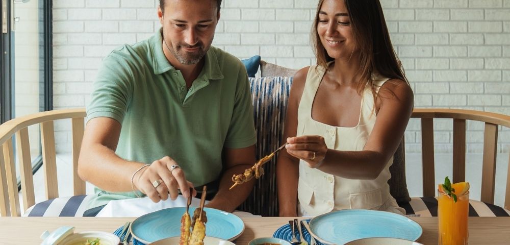 A man and a woman are seated at a table, sharing a meal of skewers of grilled meat, dips, and an orange juice drink.