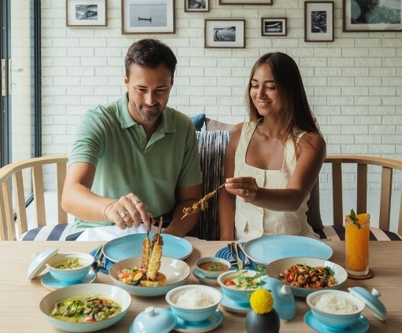 A man and a woman are seated at a table, sharing a meal of skewers of grilled meat, dips, and an orange juice drink.