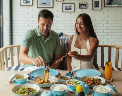 A man and a woman are seated at a table, sharing a meal of skewers of grilled meat, dips, and an orange juice drink.