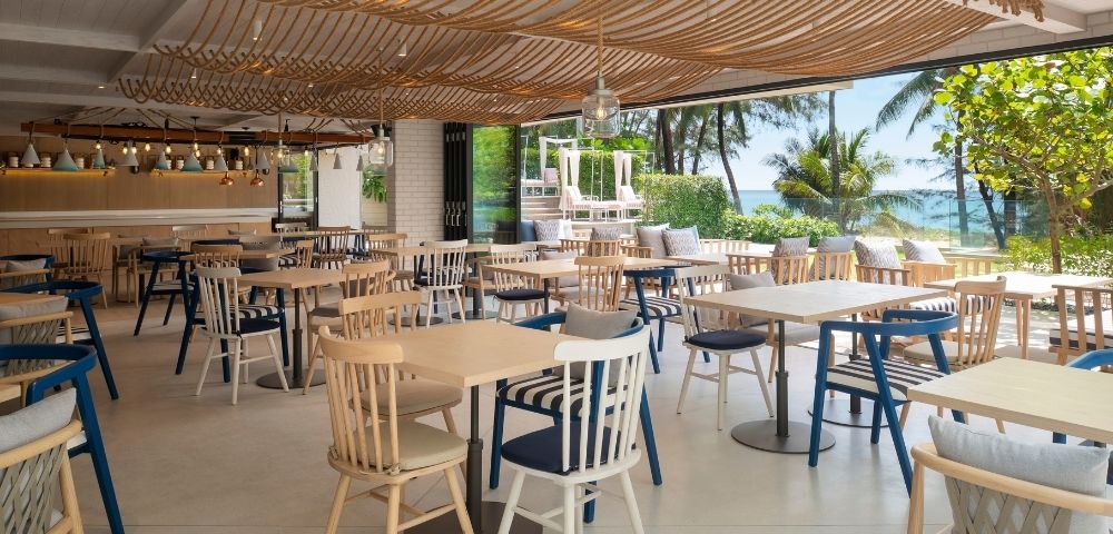 Bright beachfront restaurant with wooden tables and blue-and-white chairs, woven ceiling decor, and ocean views through open windows.