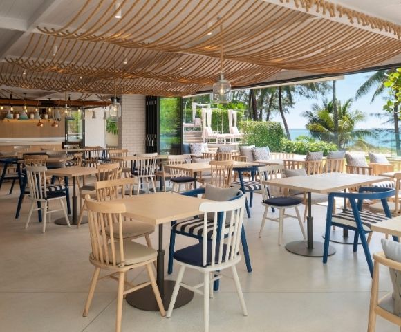 Bright beachfront restaurant with wooden tables and blue-and-white chairs, woven ceiling decor, and ocean views through open windows.