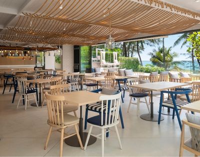 Bright beachfront restaurant with wooden tables and blue-and-white chairs, woven ceiling decor, and ocean views through open windows.