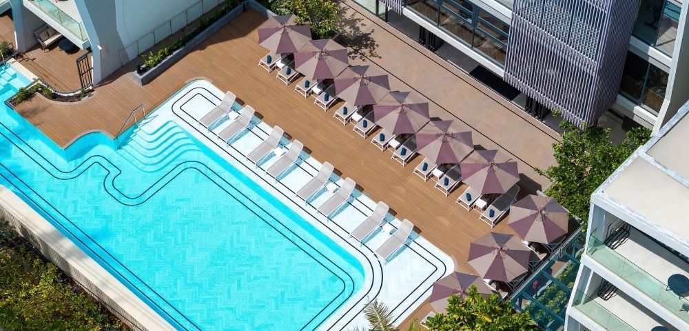 Aerial view of a bright turquoise rooftop pool lined with white loungers and mauve umbrellas beside a modern hotel, creating a calm resort feel.