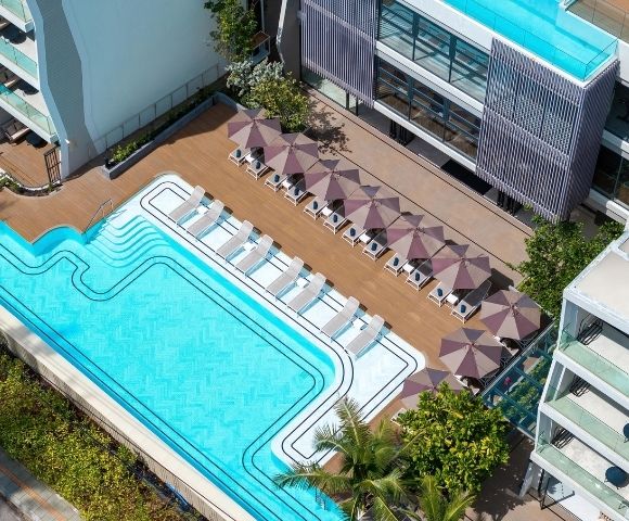 Aerial view of a bright turquoise rooftop pool lined with white loungers and mauve umbrellas beside a modern hotel, creating a calm resort feel.