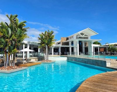 A bright blue swimming pool in the foreground with a modern white building and palm trees in the background under a clear blue sky.