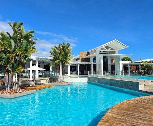 A bright blue swimming pool in the foreground with a modern white building and palm trees in the background under a clear blue sky.