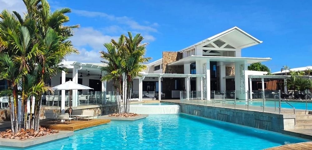 A bright blue swimming pool in the foreground with a modern white building and palm trees in the background under a clear blue sky.