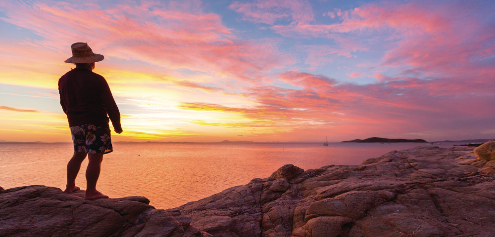 A person stands on rugged coastal rocks overlooking calm ocean waters at sunset, silhouetted against a vivid sky filled with pink, orange, and purple clouds. Warm light reflects across the sea and rocky shoreline, creating a dramatic coastal landscape with a small sailboat visible on the horizon.