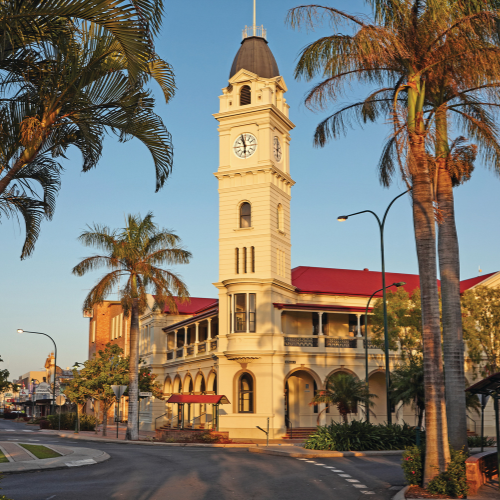A heritage-style building with a tall clock tower stands on a palm-lined street, illuminated by warm golden-hour sunlight. The cream-colored building features arched verandas and a red roof, with tropical landscaping and quiet roadways framing the civic landmark against a clear blue sky.