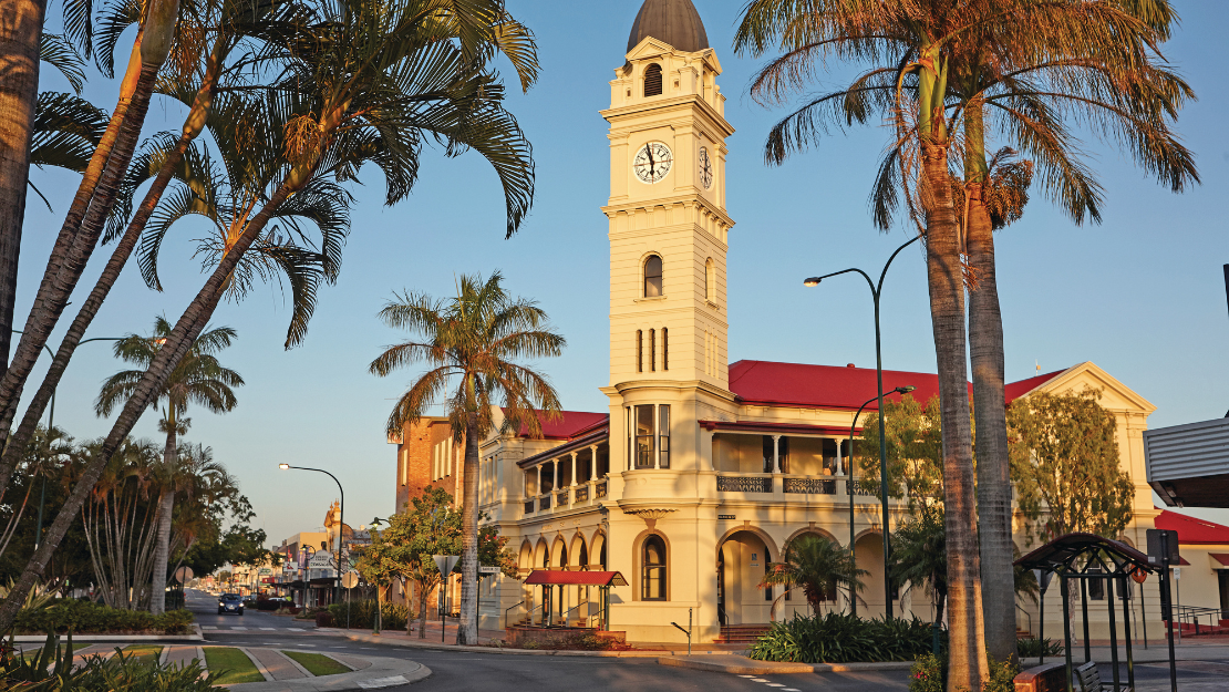 A heritage-style building with a tall clock tower stands on a palm-lined street, illuminated by warm golden-hour sunlight. The cream-colored building features arched verandas and a red roof, with tropical landscaping and quiet roadways framing the civic landmark against a clear blue sky.