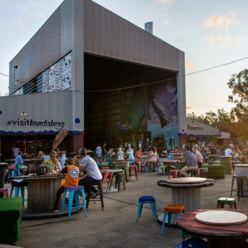 An outdoor dining and entertainment precinct is bustling with visitors seated at colorful stools and tables in an open courtyard at sunset. A large modern building featuring visible “#visitbundaberg” signage and a vibrant mural overlooks the gathering, while warm golden light filters through surrounding trees and illuminates the lively market atmosphere.