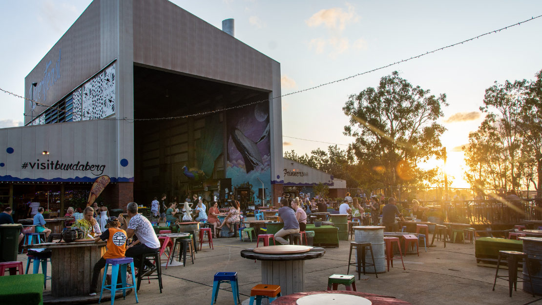 An outdoor dining and entertainment precinct is bustling with visitors seated at colorful stools and tables in an open courtyard at sunset. A large modern building featuring visible “#visitbundaberg” signage and a vibrant mural overlooks the gathering, while warm golden light filters through surrounding trees and illuminates the lively market atmosphere.