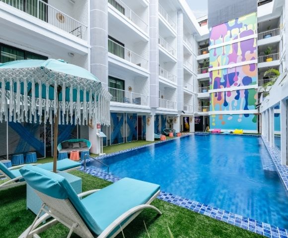 An outdoor swimming pool area with blue lounge chairs and a turquoise umbrella, surrounded by a white multi-story building with balconies and a vibrant, colorful mural on one wall.