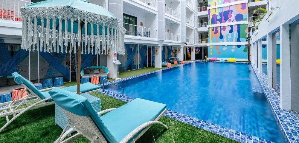An outdoor swimming pool area with blue lounge chairs and a turquoise umbrella, surrounded by a white multi-story building with balconies and a vibrant, colorful mural on one wall.