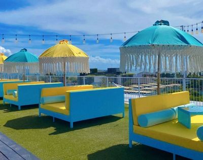 Rooftop lounge area with bright blue and yellow daybeds and fringed umbrellas under a clear blue sky with decorative lights.