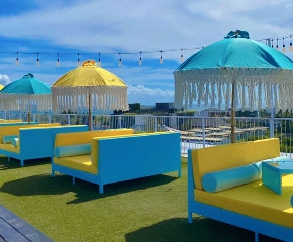 Rooftop lounge area with bright blue and yellow daybeds and fringed umbrellas under a clear blue sky with decorative lights.