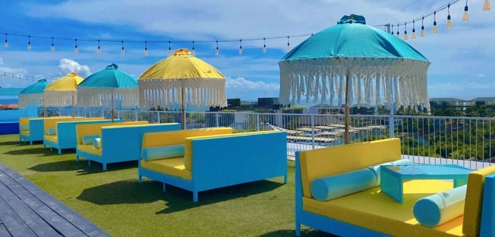 Rooftop lounge area with bright blue and yellow daybeds and fringed umbrellas under a clear blue sky with decorative lights.