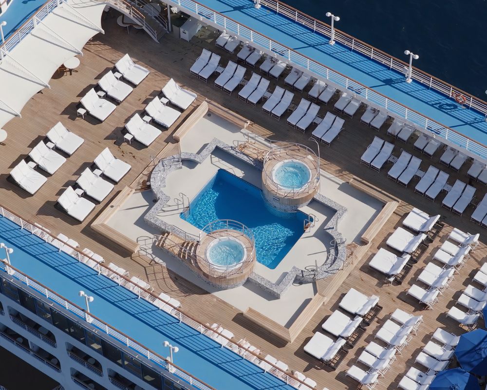 Aerial view of a cruise ship deck featuring a swimming pool with two circular hot tubs, surrounded by numerous lounge chairs and several blue umbrellas.