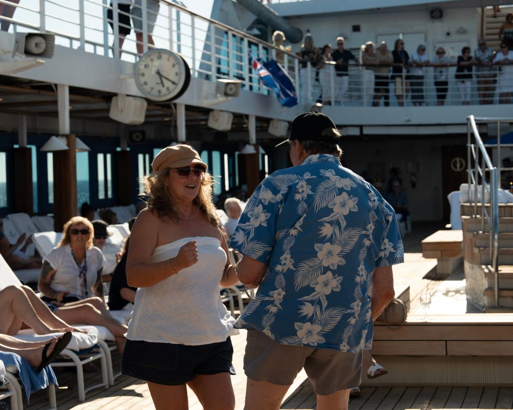 A woman with blonde curly hair wearing sunglasses and a tan newsboy cap smiles and holds her arms out as she dances on the deck of a cruise ship. She wears a white strapless top. Facing away from the camera, a man in a blue Hawaiian shirt with white flowers dances with her. In the background, several people relax on lounge chairs near the ocean.