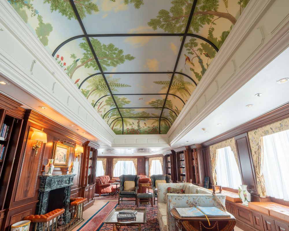 Luxurious interior lounge with floor-to-ceiling bookshelves, a fireplace, comfortable seating, and an elaborately painted vaulted ceiling depicting a tropical landscape with trees, birds, and sky.