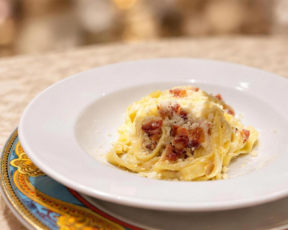 A close-up shot of fettuccine carbonara served in a white bowl. The pasta is topped with diced pancetta and grated Parmesan cheese. The bowl sits on a decorative plate with a gold and blue pattern, placed on a beige tablecloth.