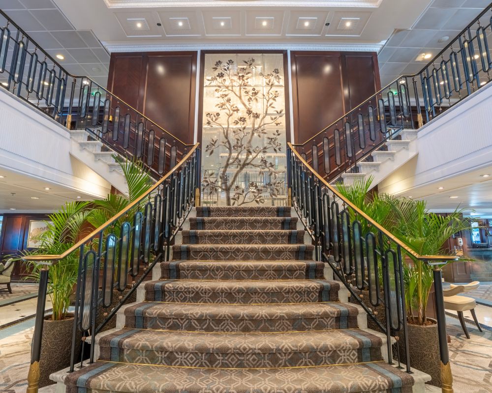 An ornate, multi-story staircase with plush patterned carpeting and dark metal railings ascends towards a large, decorative mirror featuring a stylized tree. Tall green potted plants flank the bottom steps, and warm lighting illuminates the elegant interior of what appears to be a cruise ship or hotel lobby.