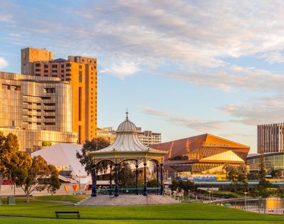 Landscaped riverside park in Adelaide featuring a decorative pavilion, green lawns, contemporary buildings, and a golden sunset sky reflected on the water.