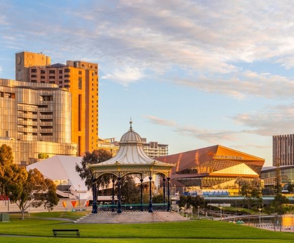 Landscaped riverside park in Adelaide featuring a decorative pavilion, green lawns, contemporary buildings, and a golden sunset sky reflected on the water.