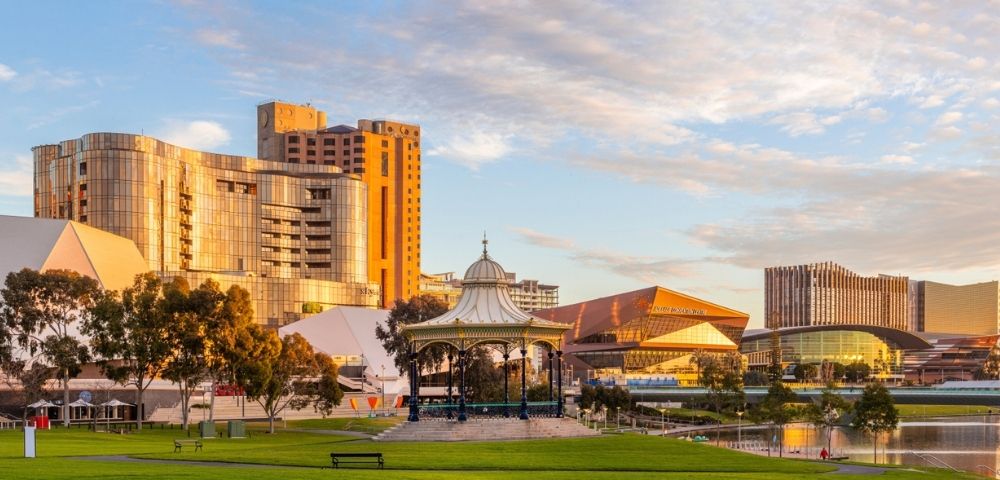 Landscaped riverside park in Adelaide featuring a decorative pavilion, green lawns, contemporary buildings, and a golden sunset sky reflected on the water.
