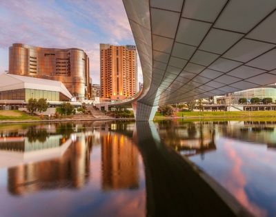 Modern Adelaide city skyline viewed from beneath a sweeping bridge, with high-rise buildings and warm sunset reflections mirrored on the calm river below.
