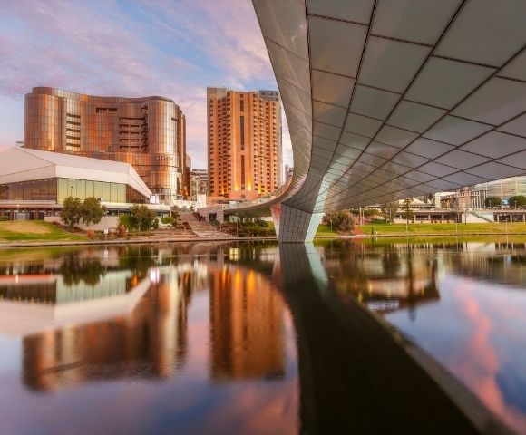 Modern Adelaide city skyline viewed from beneath a sweeping bridge, with high-rise buildings and warm sunset reflections mirrored on the calm river below.