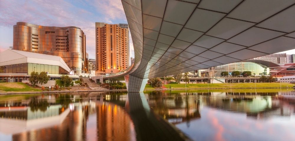 Modern Adelaide city skyline viewed from beneath a sweeping bridge, with high-rise buildings and warm sunset reflections mirrored on the calm river below.