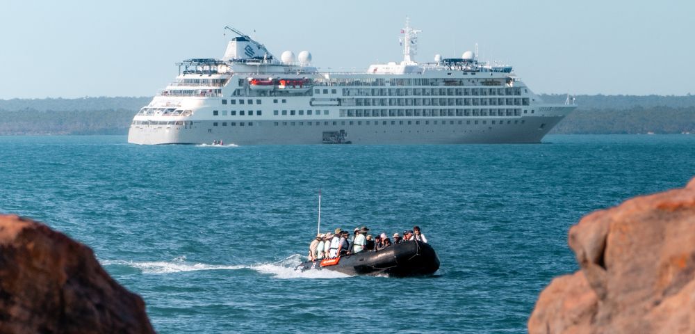 Zodiac boat with passengers approaches a large white cruise ship in the ocean, with rocky outcroppings in the foreground.
