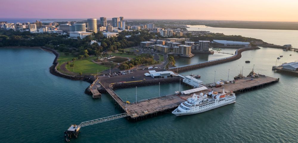 Aerial view of a cruise ship docked at a pier with a city skyline in the background at sunrise.
