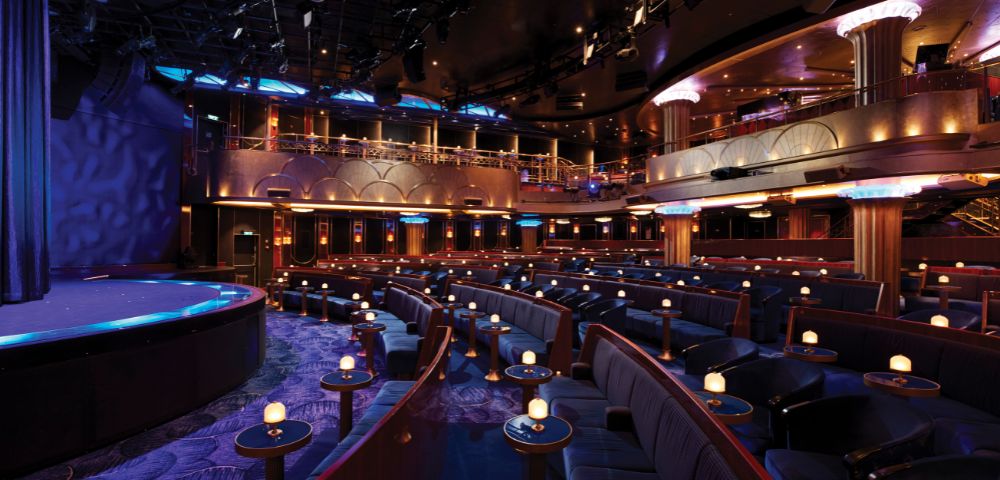An empty theater with rows of plush blue seating, small round tables with candles, a stage, and a balcony level, illuminated by dramatic stage lighting.