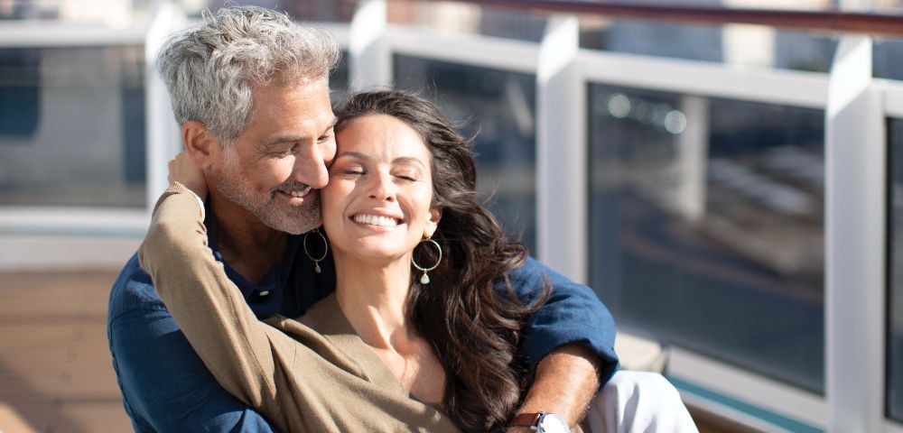 A smiling couple embraces on a cruise ship deck, with a city skyline blurred in the background under a bright sky.