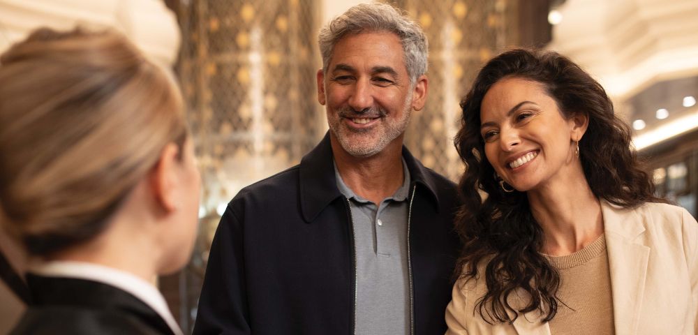 A couple smiles as a server offers them champagne flutes in an upscale interior.