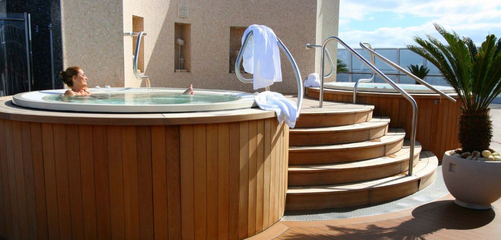 Outdoor jacuzzi with wooden exterior and curved steps, featuring a person relaxing in the water and white towels draped over railings under a sunny sky.