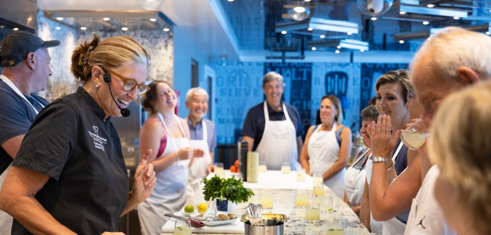 A cooking instructor in an apron and headset gestures animatedly to a group of participants in aprons, who are laughing and engaged in a cooking class.