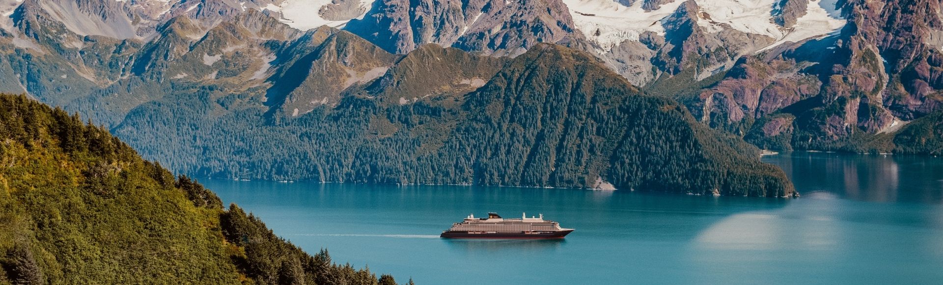 A sleek, modern cruise ship navigates through calm, turquoise waters in a majestic fjord surrounded by lush, green forested slopes and snow-capped, rugged mountains under a clear blue sky.