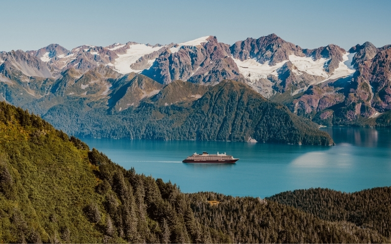 A sleek, modern cruise ship navigates through calm, turquoise waters in a majestic fjord surrounded by lush, green forested slopes and snow-capped, rugged mountains under a clear blue sky.