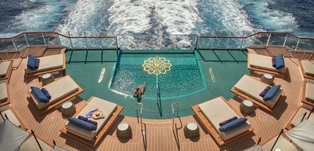 Aerial view of a person relaxing in a turquoise pool with a golden lotus design on a cruise ship deck, surrounded by lounge chairs and the ocean wake.