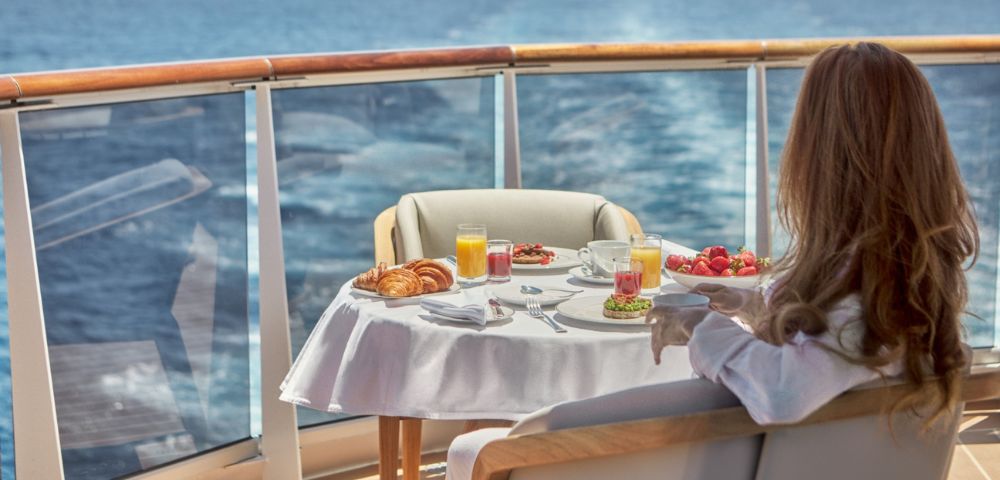 A person sits on a cruise ship balcony, facing away from the camera, overlooking the ocean while enjoying a breakfast spread of croissants, strawberries, juice, and coffee.