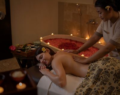 A woman reclines on a massage table receiving a back massage from a therapist, with a flower in her hair. A bathtub filled with red rose petals and illuminated by candles is visible in the background, suggesting a romantic or luxurious spa setting.